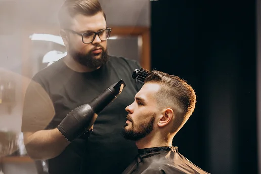 Een kapper met een bril en baard droogt en stylt het haar van een zittende klant met een föhn en borstel.