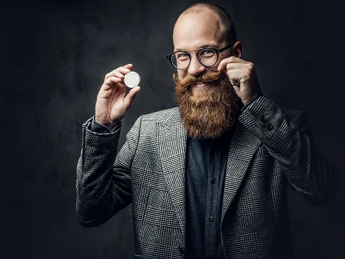 Een man met een volle rode baard en bril, gekleed in een geruit colbert, houdt glimlachend een klein rond blikje vast en trekt aan zijn snor tegen een donkere studio-achtergrond.