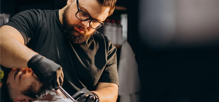 Een geconcentreerde kapper met zwarte handschoenen scheert de baard van een klant met een traditioneel open scheermes.